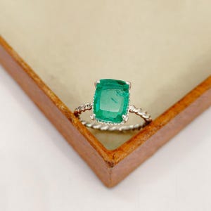 May include: A square-cut emerald ring with a twisted silver band. The vivid green gemstone is set in a silver bezel. The ring is displayed in a wooden jewellery box, showcasing its elegant design. A beautiful piece of jewellery.