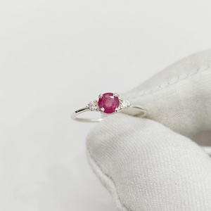 May include: A silver ring featuring a large oval ruby gemstone as the centerpiece, flanked by two smaller, pear-shaped white gemstones. The ruby is a vibrant red color, and the ring is set against a white background.