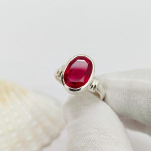 Könnte beinhalten: Ein silberner Ring mit einem großen, oval geschliffenen Rubin. Der Rubin ist tiefrot und in eine polierte Silberschiene mit dekorativen Seitenakzenten gefasst. Der Ring wird vor weißem Hintergrund gezeigt. Ein Schmuckstück.