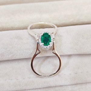 Puede incluir: Un anillo de plata con una gran piedra ovalada de color verde esmeralda rodeada por un halo de piedras pequeñas transparentes.