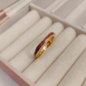 May include: A gold-toned ring featuring a thin, recessed channel filled with a textured, deep red inlay. The band has a smooth, polished finish on the edges, creating a contrast between the metallic gold surface and the dark red center detail.