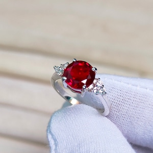 Op de afbeelding: Een zilveren ring met een grote, ronde, rode robijn-edelsteen omringd door kleinere, heldere edelstenen.