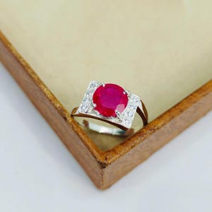 Può includere: Un anello d'argento con una grande gemma rossa rotonda e vibrante come punto focale. La gemma è circondata da un'incastonatura quadrata ornata da pietre più piccole e chiare. L'anello è posto su uno sfondo marrone chiaro.
