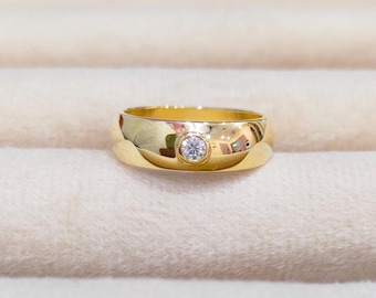 Puede incluir: Un anillo de banda de oro con una sola piedra preciosa redonda y transparente engastada en el centro.