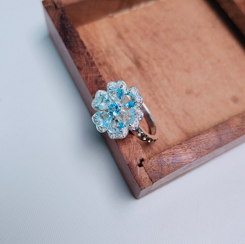 May include: A silver ring with a blue topaz flower design. The ring is set in a wooden box.