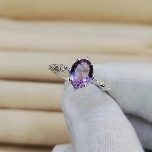 May include: A silver ring featuring an oval-cut, faceted purple amethyst gemstone. The band is adorned with small, clear accent stones. The ring is displayed against a soft, neutral background.