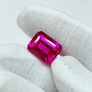 May include: A vibrant pink gemstone with a rectangular emerald cut. The gemstone is held in a gloved hand.