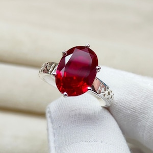 May include: A silver ring with a large, oval-shaped ruby gemstone. The ring has a delicate, intricate design.