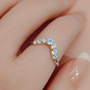 以下が含まれることがあります： 銀色の曲線的なリングで、6つの丸い虹色のムーンストーンが並んでいます。バンドはV字型の輪郭を描いており、石のサイズはわずかに異なり、表面には柔らかな青い光の反射が見られます。