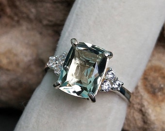 May include: A silver ring featuring a large, light green gemstone and smaller clear gemstones on the band.