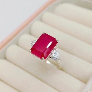 May include: A silver ring with a large, rectangular, deep red gemstone. Two smaller, triangular, clear gemstones are either side of the main stone. The ring is set against a light-coloured, padded jewellery box.