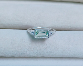 May include: A silver ring with a light green rectangular gemstone and two smaller clear gemstones on the band.