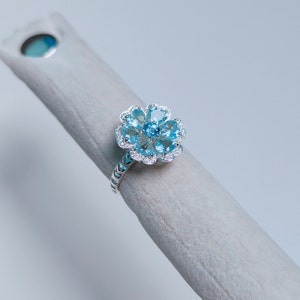 May include: A silver ring with a blue topaz flower design. The ring has a textured band and a flower-shaped setting with multiple blue topaz stones.