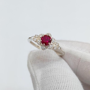Könnte beinhalten: Ein silberner Ring mit einem roten Rubin-Edelstein, der von kleineren weißen Edelsteinen umgeben ist.