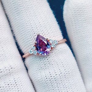 以下が含まれることがあります： ローズゴールドのバンドに、洋梨型の紫色の宝石と、より小さな透明な宝石がセットされたリングです。