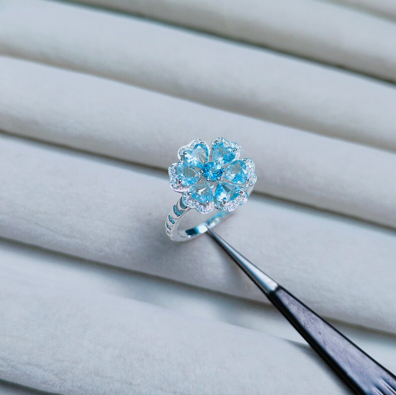 May include: A silver ring with a flower-shaped design featuring blue gemstones. The ring is set on a white background.