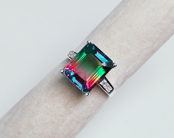 May include: A silver ring with a large, emerald-cut gemstone that is green, blue, and pink. The gemstone is surrounded by smaller, clear gemstones.