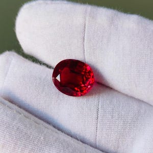 Peut inclure: Une pierre précieuse rubis de forme ovale et de couleur rouge profond. La pierre est présentée sur un fond blanc, mettant en valeur ses facettes et sa brillance. Un exemple frappant de joaillerie.