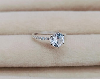 Puede incluir: Un anillo de plata con una piedra preciosa grande, redonda y transparente rodeada de piedras preciosas más pequeñas y transparentes.