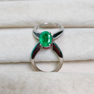 Może przedstawiać: Srebrny pierścionek z żywym, zielonym owalnym kamieniem szlachetnym. Pierścionek ma unikalny, otwarty design, z obrączką zakrzywioną wokół klejnotu. Jasnozielony kamień jest centralnym punktem na neutralnym tle.