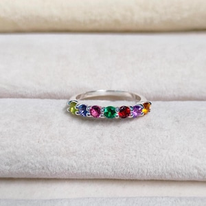 Puede incluir: Un anillo de banda de plata con una fila de piedras preciosas de colores. Las piedras son verdes, azules, rosas, rojas, moradas y naranjas.