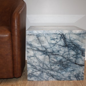 May include: A square side table with a marble-like pattern in shades of gray and blue. The table is positioned next to a brown leather armchair. The table's design features dark blue veining against a light gray background, creating a natural stone appearance.