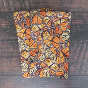May include: Folded fabric with a vibrant pattern of monarch butterfly wings in shades of orange, yellow, black, and white. The fabric is rectangular and appears to be a scarf or a similar accessory. The background is a dark brown wooden surface.