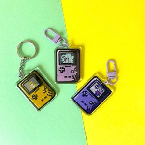 May include: Three keychains featuring pixelated game console designs. The keychains are purple, yellow, and blue. Each keychain has a different game console design.