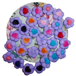 May include: A collection of purple fabric flowers with various colored felt hearts in the center. The flowers are arranged on a white and green patterned surface.