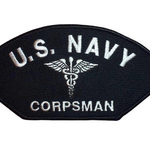 Może przedstawiać: Czarna naszywka z białym haftowanym tekstem: "U.S. NAVY CORPSMAN." Symbol medyczny kaduceusza jest wyśrodkowany między "NAVY" i "CORPSMAN." Naszywka ma unikalny kształt wachlarza.
