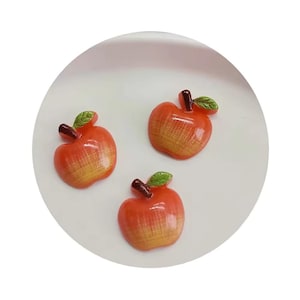 May include: Three red and yellow apple shaped resin cabochons with green leaves. The cabochons are on a white plate.