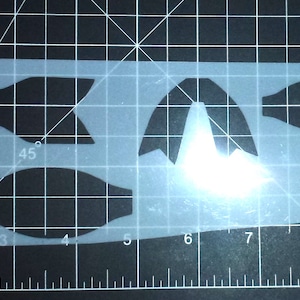 May include: A white plastic stencil with two bow tie shapes, one measuring 60 degrees and the other 45 degrees. The stencil is placed on a black gridded surface.