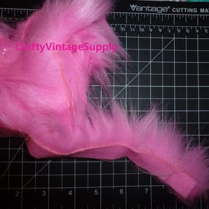 May include: A pink faux fur trim with a slight sheen. The trim is approximately 10 inches long and is shown on a black cutting mat with white grid lines.
