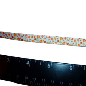 May include: White ribbon with a repeating pattern of orange and brown pumpkins. The ribbon is measured against a black ruler with inch markings.