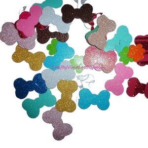 Peut inclure: Assortiment de formes d'os et de nœuds recouverts de paillettes dans différentes couleurs, notamment rose, bleu, vert, or, argent et marron. Les formes sont éparpillées sur un fond blanc.