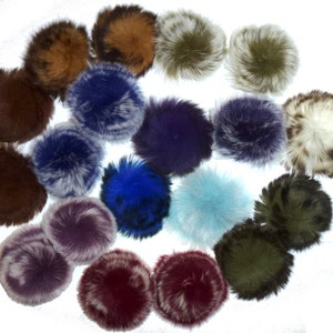 May include: A collection of fluffy pom-poms in various colors, including brown, blue, purple, and green. The pom-poms have a soft, textured appearance, suggesting they are made of fur or a similar material. They are arranged against a white background.