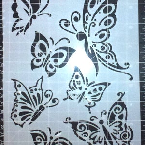 Può includere: Un foglio di stencil con sei diversi disegni di farfalle. Le farfalle sono tutte nere e hanno dettagli intricati. Il foglio di stencil è su una superficie a griglia.