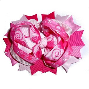 May include: A pink and white hair bow with a heart and swirl pattern. The bow is made of ribbon and has a star-shaped design.