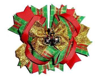 May include: A festive Christmas hair bow featuring red, green, and gold ribbons. The bow has a layered design with glitter accents and small silver bells in the center. The bow is a holiday accessory.