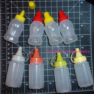 May include: A collection of clear plastic squeeze bottles with colorful caps in yellow, red, orange, and gray. The bottles are arranged on a black cutting mat with white grid lines. The image includes the text "Crafty Vintage Supply".