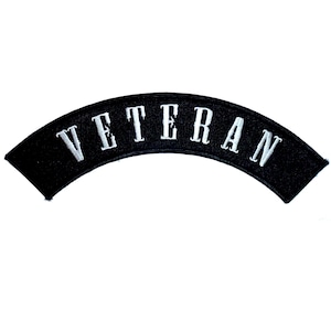 May include: A curved, black embroidered patch featuring the word VETERAN stitched in white capital letters. The patch has a distinct arc shape, designed for placement on clothing or gear, with a clean, high-contrast design and finished edges.