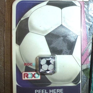 以下が含まれることがあります： 銀色の枠が付いた白黒のサッカーボールのステッカー。ステッカーは紫色の背景のカードに貼られており、白い文字で「PEEL HERE Reusable Sticker!」と書かれています。
