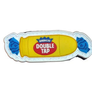Puede incluir: Un dije de zapato amarillo y rojo "American Double Tap" con detalles azules. El dije tiene forma de bala con el texto "American Double Tap" en un óvalo rojo.