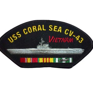 Puede incluir: Parche bordado negro con el texto "USS CORAL SEA CV-43 VIETNAM" en amarillo y rojo. Presenta una imagen detallada de un barco y cintas coloridas. El parche tiene una forma única.