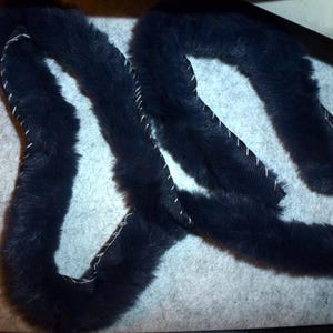 May include: A length of black faux fur trim with white stitching. The trim is sewn onto a black fabric backing. The fur is soft and fluffy, and the stitching is visible along the edges. The trim is likely for clothing or craft projects.