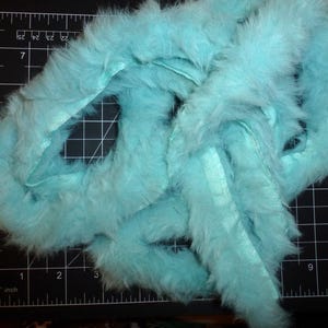 Puede incluir: Una longitud de boa de plumas azul claro y esponjosa sobre una esterilla de corte negra con medidas en pulgadas. La boa tiene una cinta de satén que la atraviesa. La boa es de un color brillante y liso.