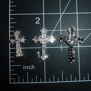 May include: Three silver cross charms with rhinestone accents. The center cross has a heart shape in the center. The cross on the right has black rhinestones.