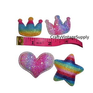 May include: Four colourful felt appliques with a rainbow glitter finish. The appliques are a heart, a star, and two crowns. The crowns are different sizes and styles. A measuring tape is included for size reference.