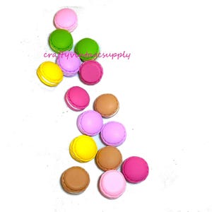 May include: Assortment of colorful macarons, including pink, yellow, green, brown, and purple. The macarons are arranged in a scattered pattern on a white background.