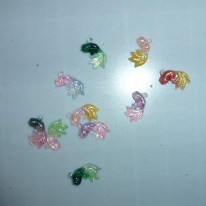 May include: A collection of small, colorful fish tail charms in various colors, including pink, yellow, green, and red. Each charm has a small loop for attaching to jewelry or other crafts. The charms are scattered on a white surface.